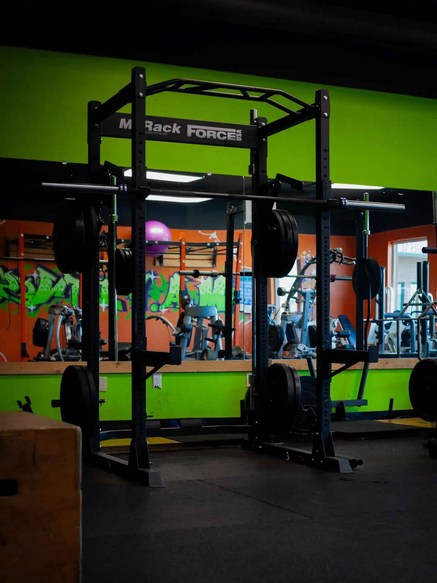 The interior of Mission Accomplish Gym in Kennewick showing a Force USA power rack, mirrors, orange and green walls, and turf flooring — the training floor Hands of Time Wellness works out of