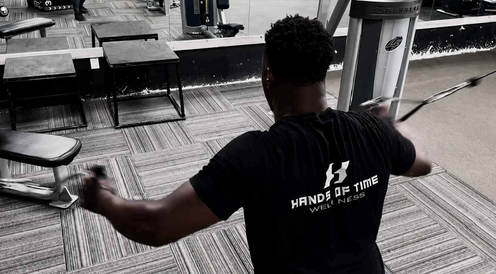 Byron Laws in a Hands of Time Wellness t-shirt performing a standing cable row exercise on the Mission Accomplish gym floor