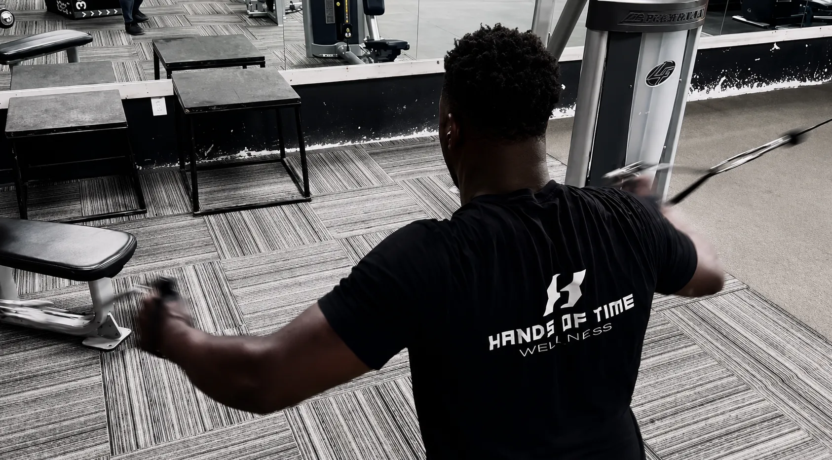 Byron Laws in a Hands of Time Wellness t-shirt performing a standing cable row exercise on the Mission Accomplish gym floor